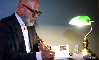 Ein Mann sitzt links im Bild, er trägt Anzug und eine schwarze Hornbrille und Bart. Er schreibt etwas an einem Schreibtisch. Auf dem Tisch steht eine kleine grüne Lampe.