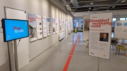 Heller Ausstellungsraum mit Infoplakaten an weißen Wänden, roter Leitlinie am Boden, Bildschirm „Wege für alle“ links und Tafel „Düsseldorf nachhaltig mobil“ rechts.