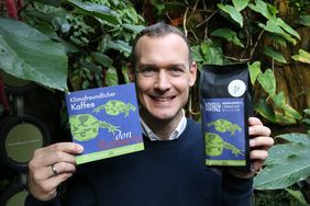 Aquazoodirektor Dr. Jochen Reiter präsentiert eine Packung des Aquazoo-Kaffees und hält einen Flyer zur Kooperation in der Hand. Im Hintergrund sind die Pflanzen der Tropenhalle des Aquazoo zu sehen.