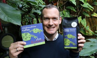 Aquazoodirektor Dr. Jochen Reiter präsentiert eine Packung des Aquazoo-Kaffees und hält einen Flyer zur Kooperation in der Hand. Im Hintergrund sind die Pflanzen der Tropenhalle des Aquazoo zu sehen.