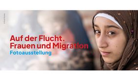 Auf dem Bild ist, im Vordergrund stehend, eine Frau zu sehen. Im unscharfen Hintergrund ist ein Kleinkind in den Armen einer Frau erkennbar. Links im Bild ist der Schriftzug: "Auf der Flucht. Frauen und Migration" Fotoausstellung lesbar. © Erol Gurian