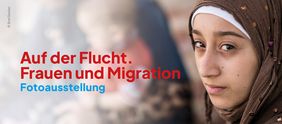 Auf dem Bild ist, im Vordergrund stehend, eine Frau zu sehen. Im unscharfen Hintergrund ist ein Kleinkind in den Armen einer Frau erkennbar. Links im Bild ist der Schriftzug: "Auf der Flucht. Frauen und Migration" Fotoausstellung lesbar. © Erol Gurian