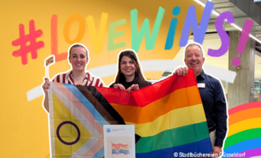 Foto mit drei Menschen in der Zentralbibliothek, die eine Regenbogenflagge in die Kamera halten und der Schriftzug: "#Lovewins"