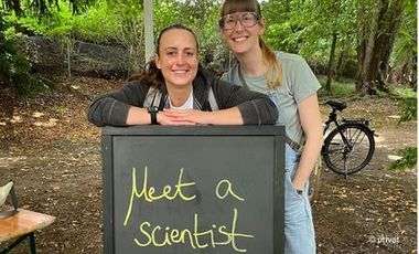 Zwei Personen stehen hinter einer Kreideklapptafel, auf der "Meet a Scientist" steht. Im Hintergrund ist Wald und ein Fahrrad zu sehen