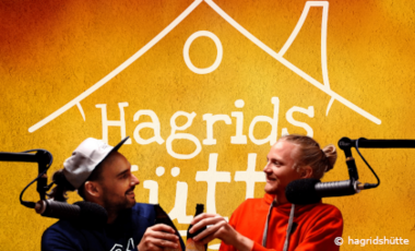 Foto von zwei jungen Männern, die einen Podcast aufnehmen. Im Hintergrund steht der Schriftzug "Hadrids Hütte" auf der Wand.