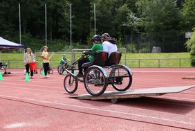 Spezial Fahrrad mit drei Rädern mit zwei Radfahrenden, die über eine Wippe fahren die als Hindernis auf einem Parcours aufgebaut ist. Im Hintergrund stehen 2 freiwillige Helfer.