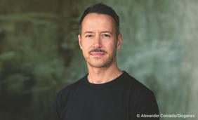 Profilfoto eines Mannes, der im Dreiviertelprofil zur Kamera steht. Er trägt ein schwarzes T-Shirt und steht vor grünem verwaschenem Hintergrund