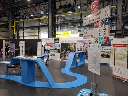 Messestand zur Radmobilität in Düsseldorf mit Infotafeln, Karten und Wegweisern; blaues, geschwungenes Display im Vordergrund, Fahrräder und Besucher im Hintergrund.